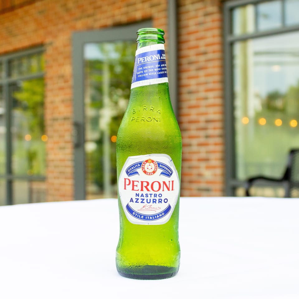 Chilled green Peroni Nastro Azzurro beer bottle on a white table, with a brick-walled restaurant visible behind it.