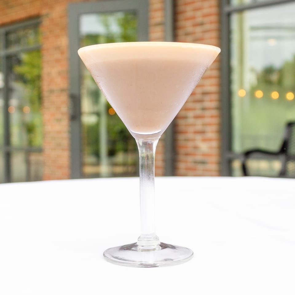 Frosted pale pink cocktail in a martini glass on a white table, with a brick-walled restaurant patio visible behind it.