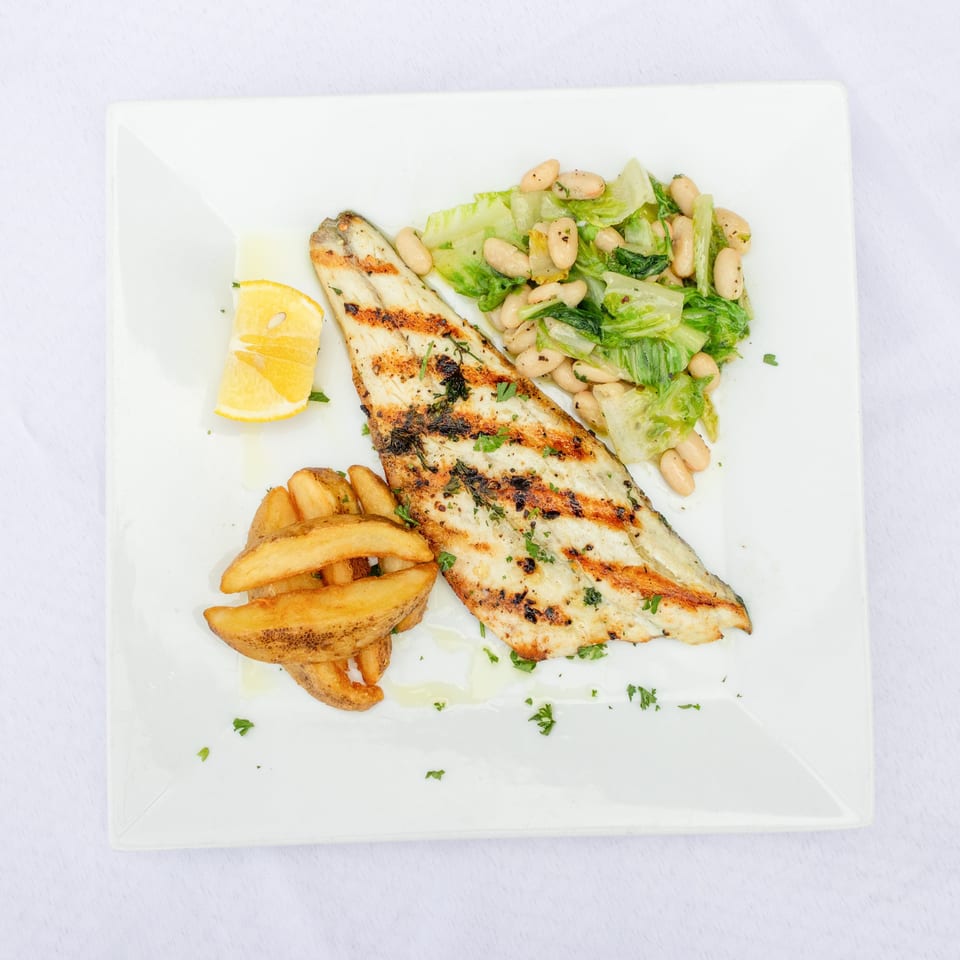 Grilled fish fillets served with potato wedges, lemon, and a white bean and lettuce salad on a square plate.