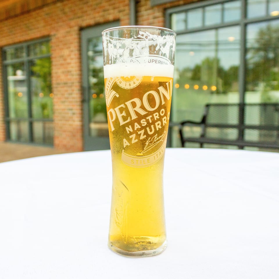 Tall glass of golden Peroni beer with a foamy head on a white table outside a brick restaurant.