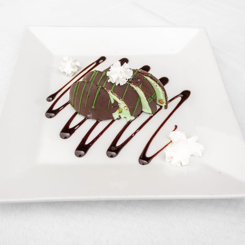 Chocolate dessert with mint-green filling, chocolate and green drizzle, whipped cream, and nuts on a white plate.