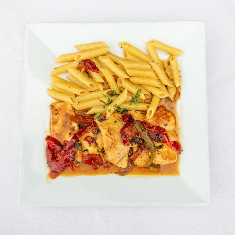 Penne pasta and sliced chicken topped with sun-dried tomatoes and herbs in a creamy sauce on a white plate.