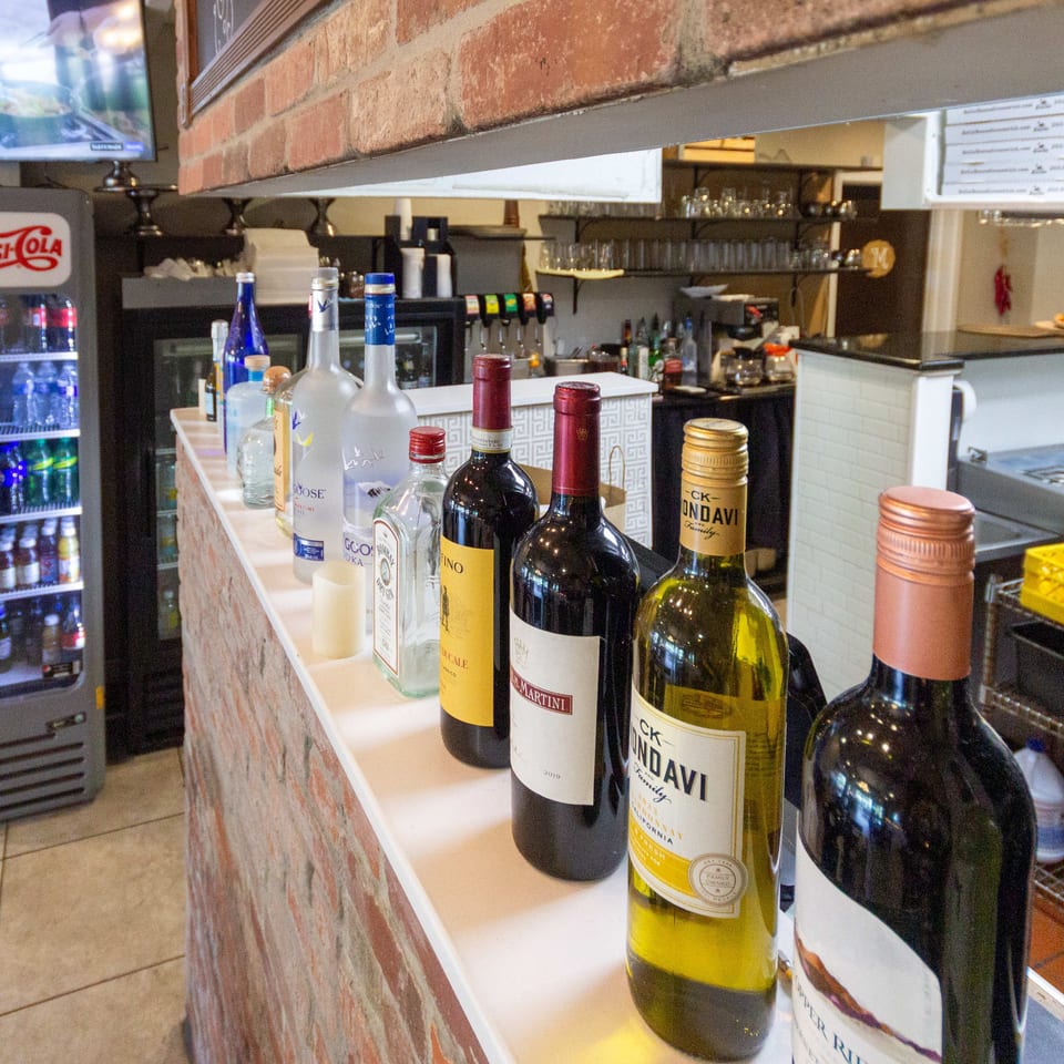 Restaurant bar counter displaying wine bottles, beverage refrigerator, drink taps, glassware, and food service equipment against a brick wall.