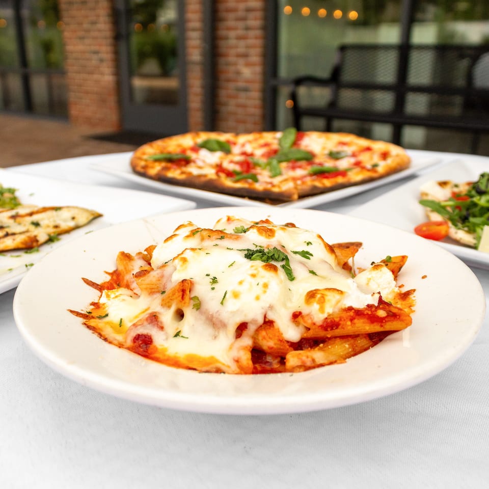 Italian restaurant spread featuring cheesy baked pasta, margherita pizza, flatbread, and salad on a white-tablecloth table.