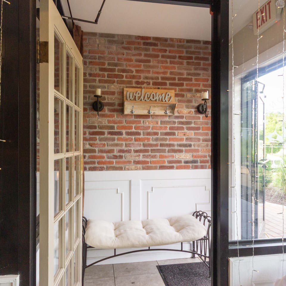 Restaurant entryway with exposed brick walls, a welcome coat rack, cushioned bench, candles, and string lights by the window.