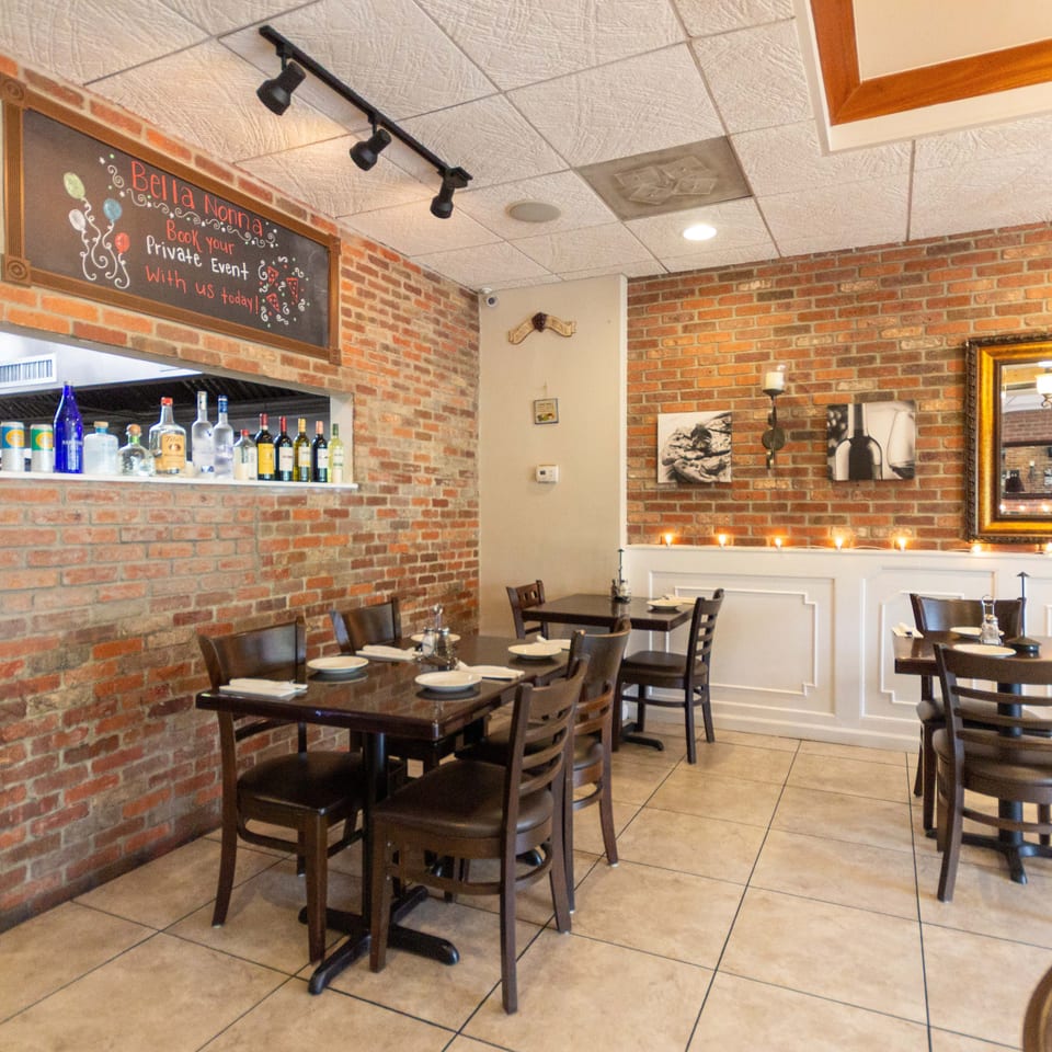 Bella Nonna restaurant dining room with brick walls, set tables, a bar shelf of bottles, candles, and framed artwork.